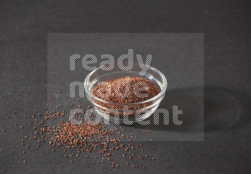 A glass bowl full of garden cress seeds surrounded by seeds on a black flooring