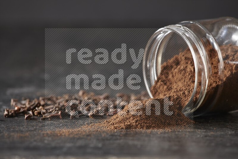 A flipped glass jar full of cloves powder with cloves spread on a textured black flooring
