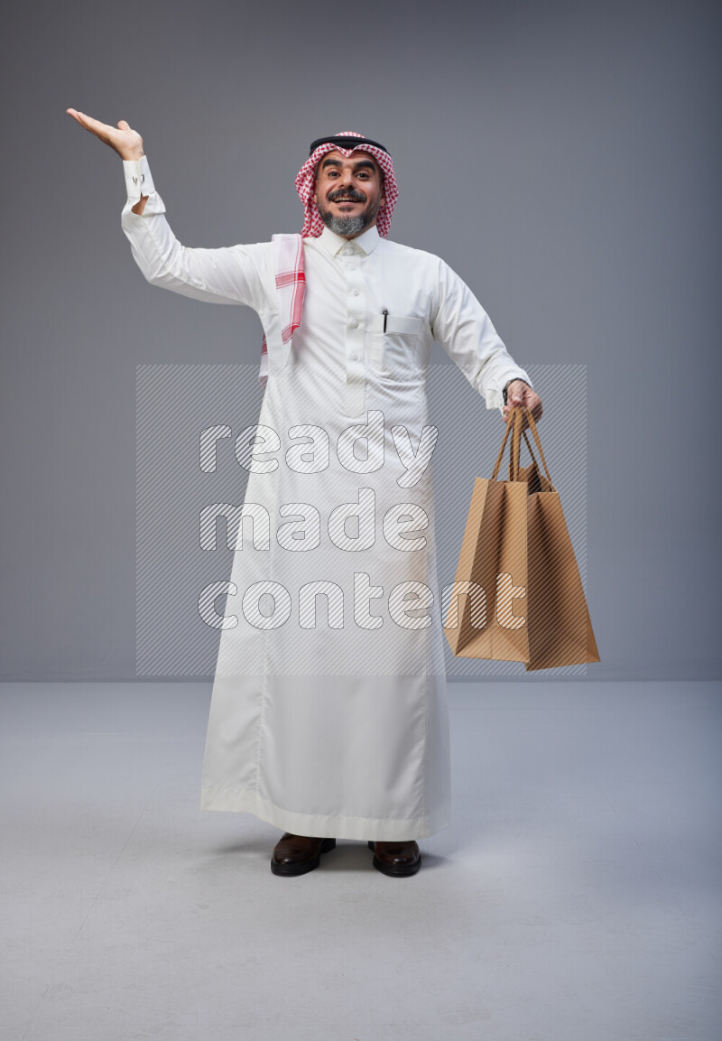 Saudi man Wearing Thob and red Shomag standing holding shopping bag on Gray background