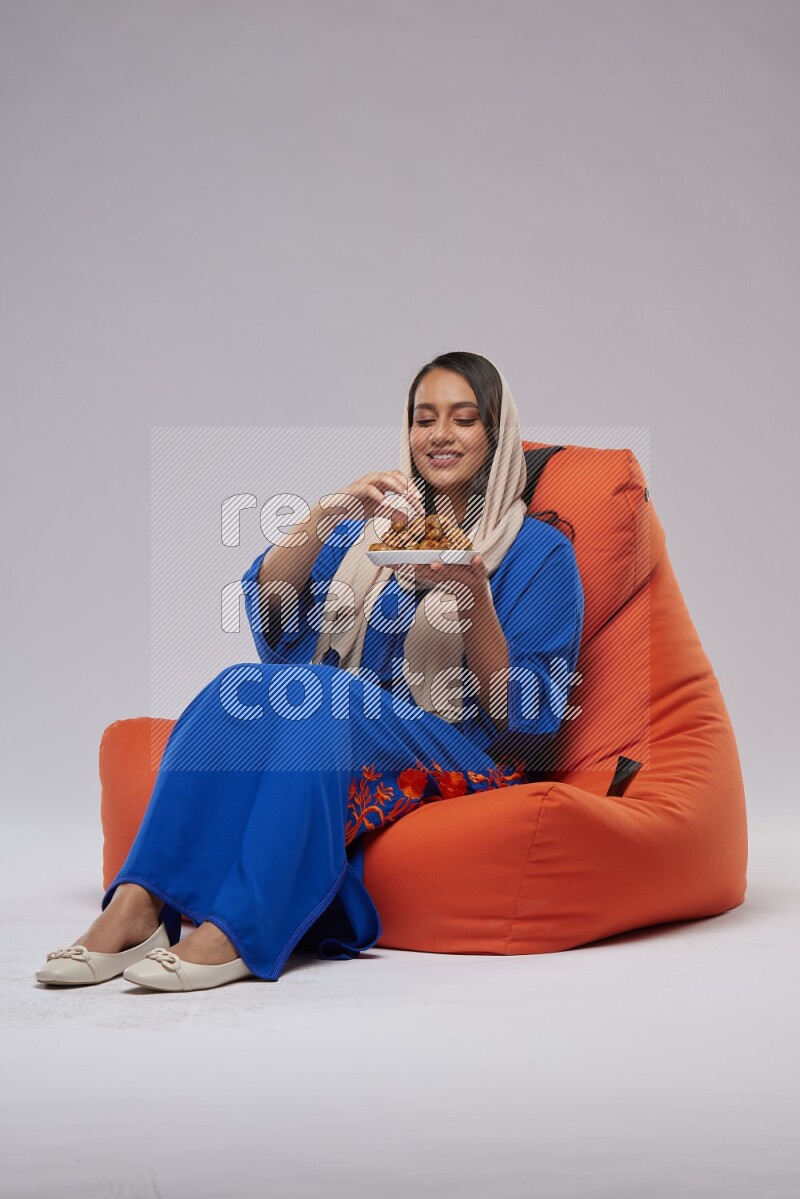 A Woman sitting on an orange beanbag wearing Jalabeya holding a plate of dates