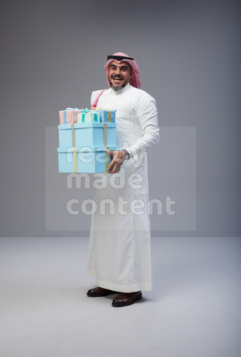 Saudi man Wearing Thob and red Shomag standing holding gift box on Gray background