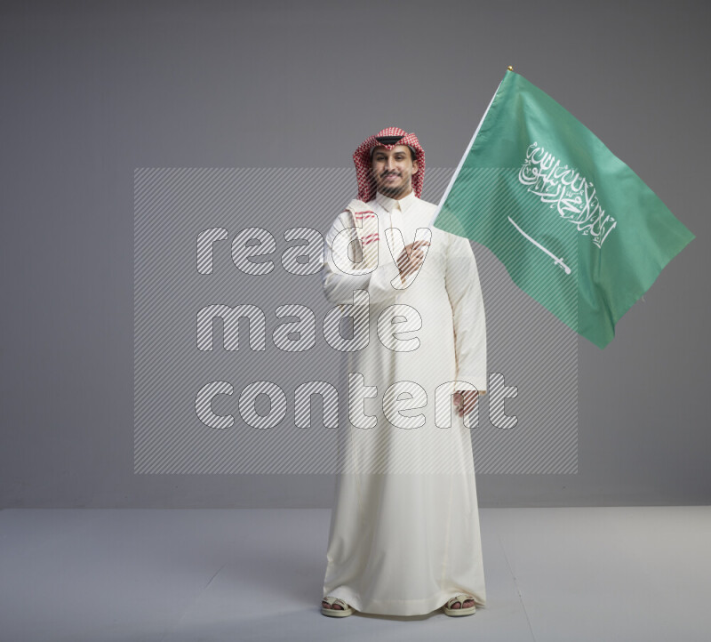 A Saudi man standing wearing thob and red shomag raising big Saudi flag on gray background