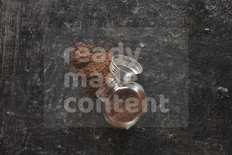 A flipped glass spice jar full of cloves powder and powder came out of it on textured black flooring
