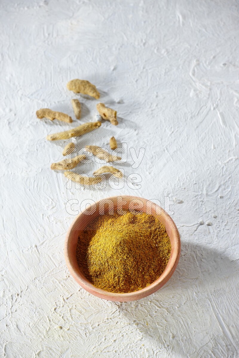 A wooden bowl full of turmeric powder with dried whole fingers on textured white flooring