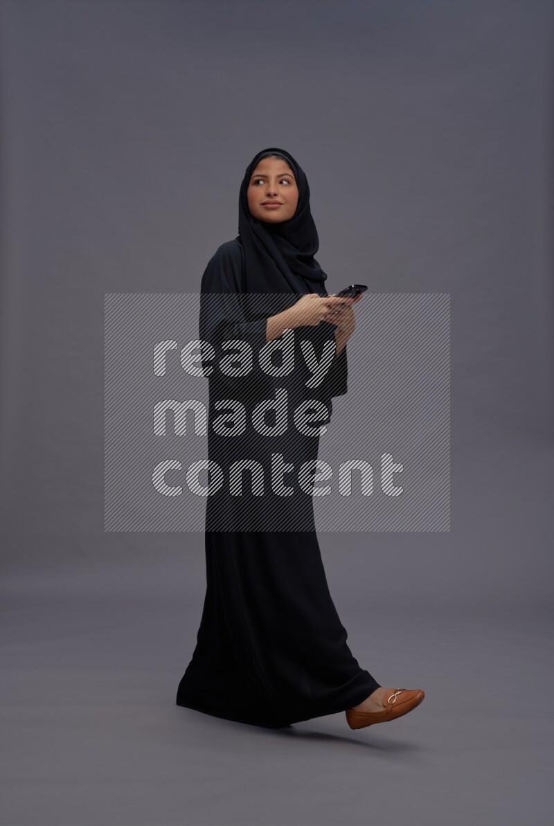 Saudi woman wearing Abaya standing texting on phone on gray background
