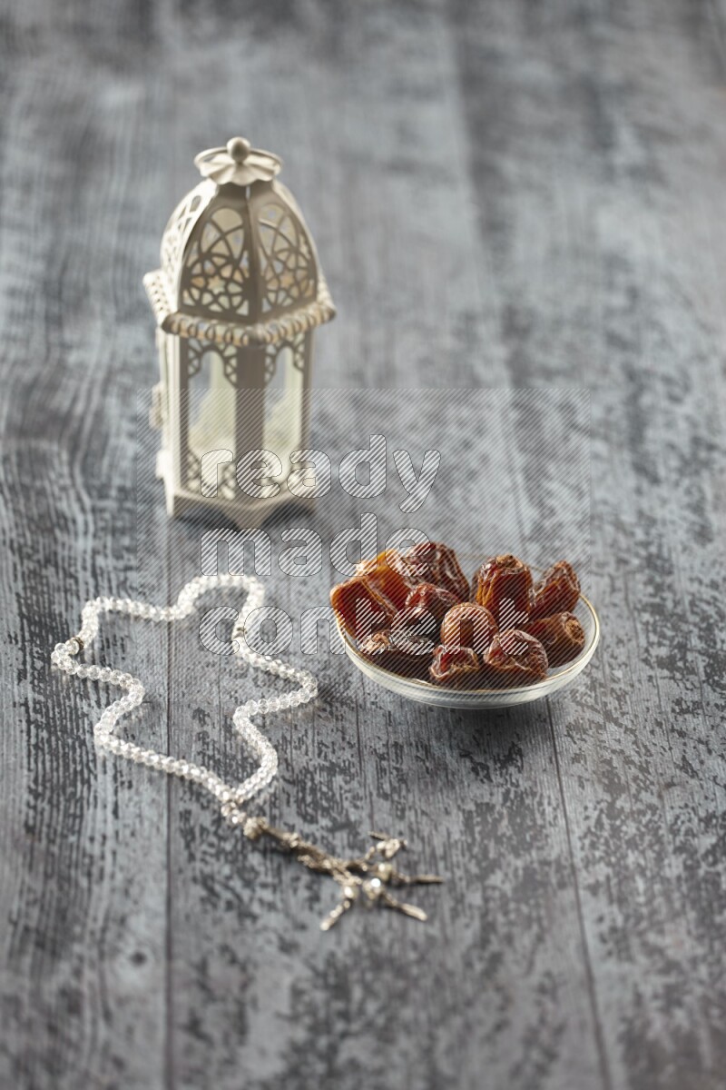A white lantern with different drinks, dates, nuts, prayer beads and quran on grey wooden background