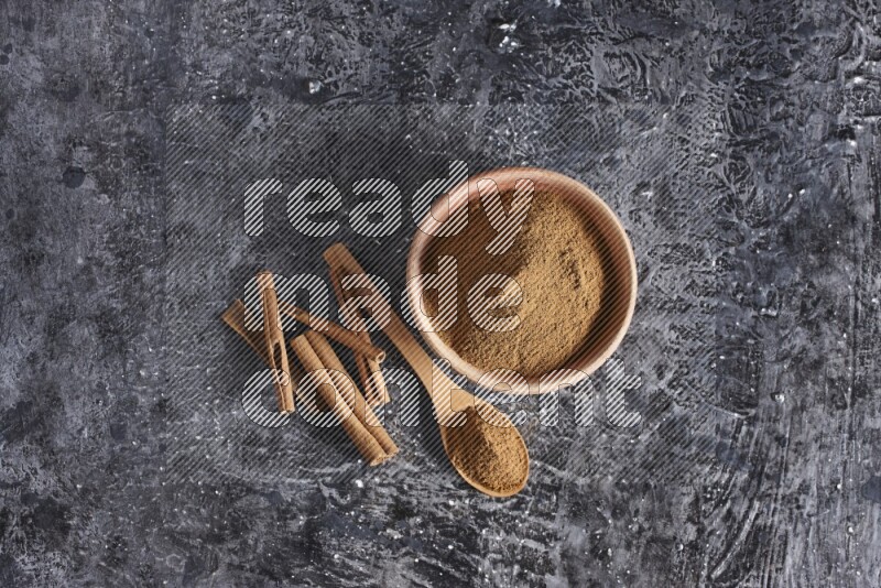 wooden bowl full of cinnamon powder and a wooden spoon full of it with cinnamon sticks on a textured black background