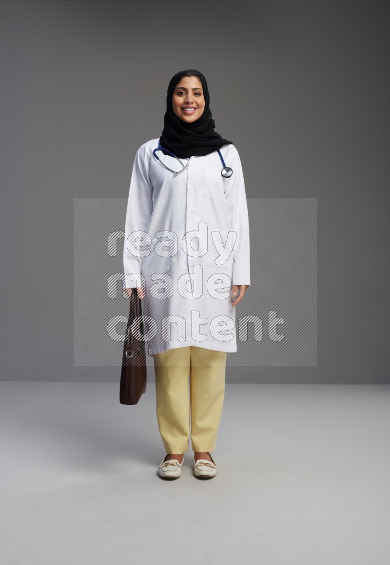Saudi woman wearing lab coat with stethoscope standing holding bag on Gray background