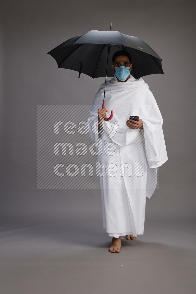 A man wearing Ehram and mask Standing holding umbrella on gray background on gray background