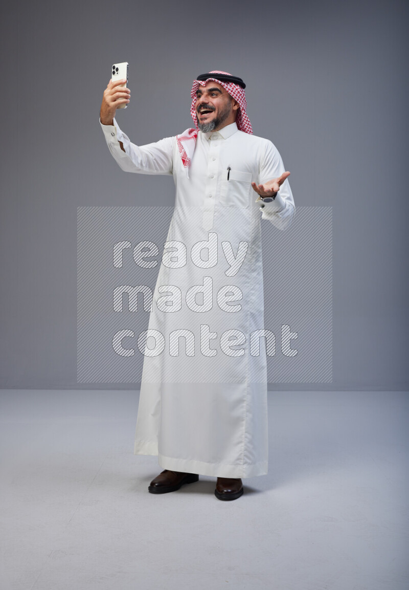 Saudi man Wearing Thob and red Shomag standing taking selfie on Gray background