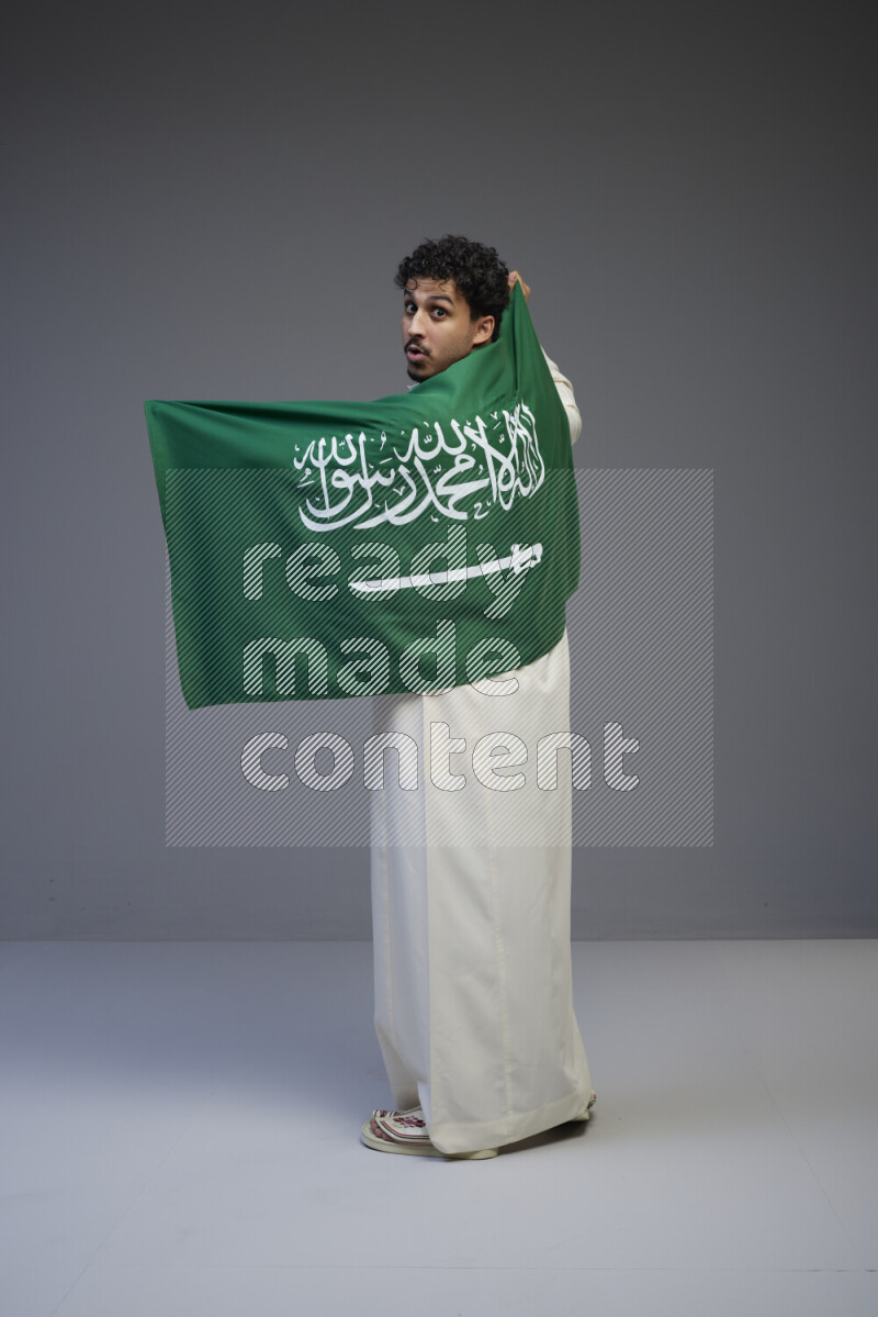 A Saudi man standing wearing thob holding big Saudi flag on gray background