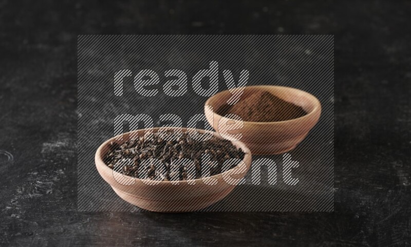 2 wooden bowls full of cloves powder and whole cloves on a textured black flooring