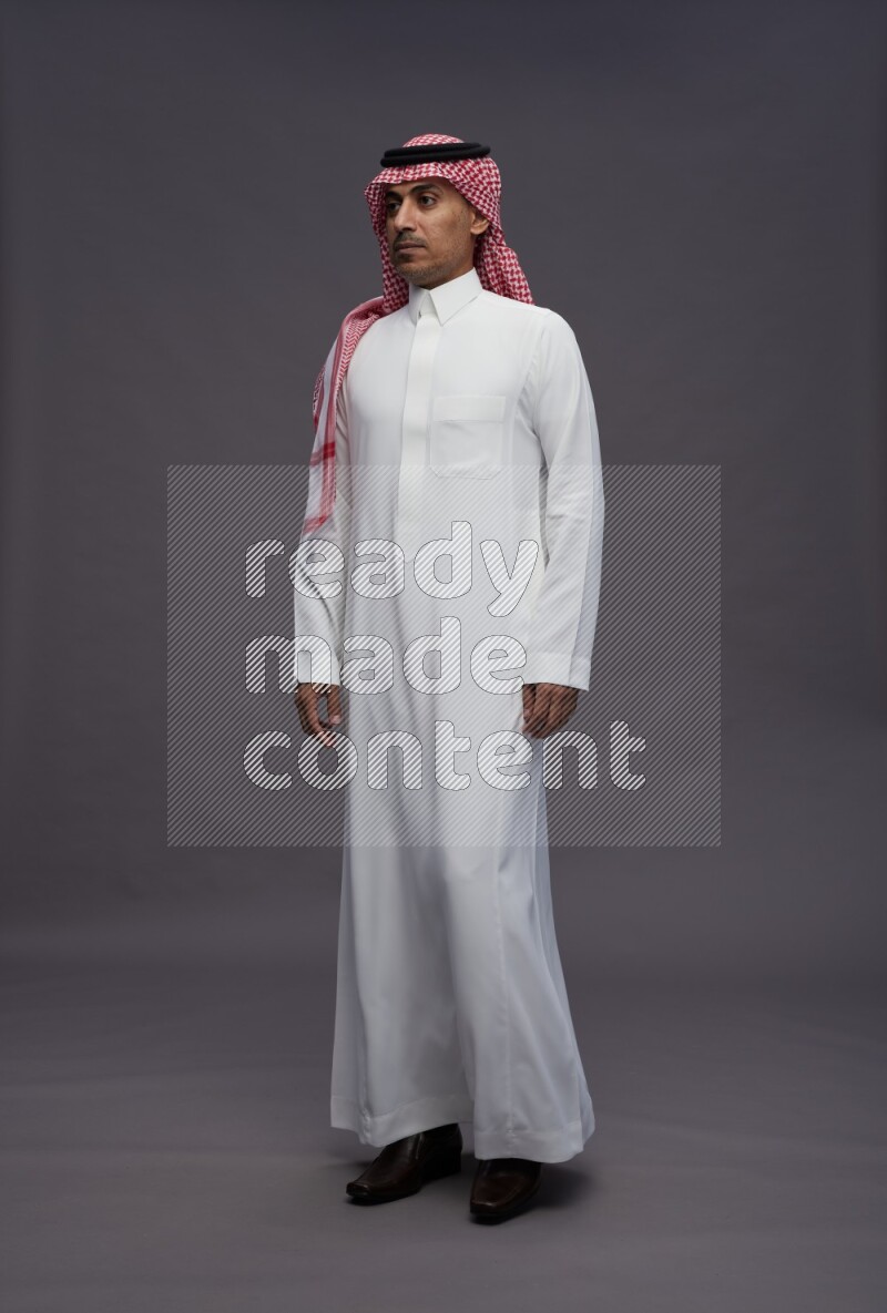 Saudi man wearing thob and shomag standing interacting with the camera on gray background