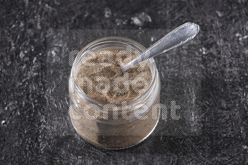 A glass jar full of black pepper powder and a metal spoon on a textured black flooring