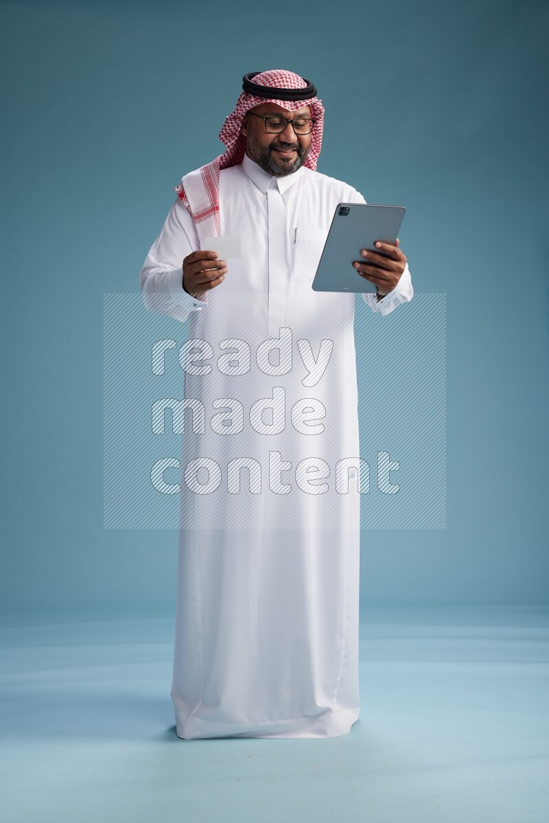 Saudi Man with shimag Standing holding ATM card while working on tablet on blue background
