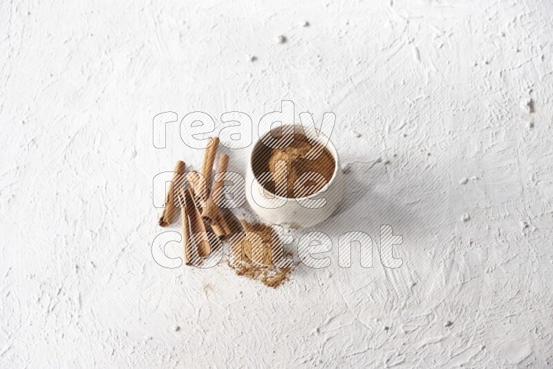 Ceramic beige bowl full of cinnamon powder and a metal spoon with cinnamon sticks next of it on a textured white background
