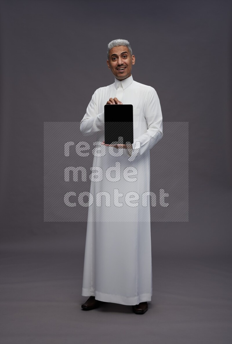 Saudi man wearing thob standing showing tablet to camera on gray background