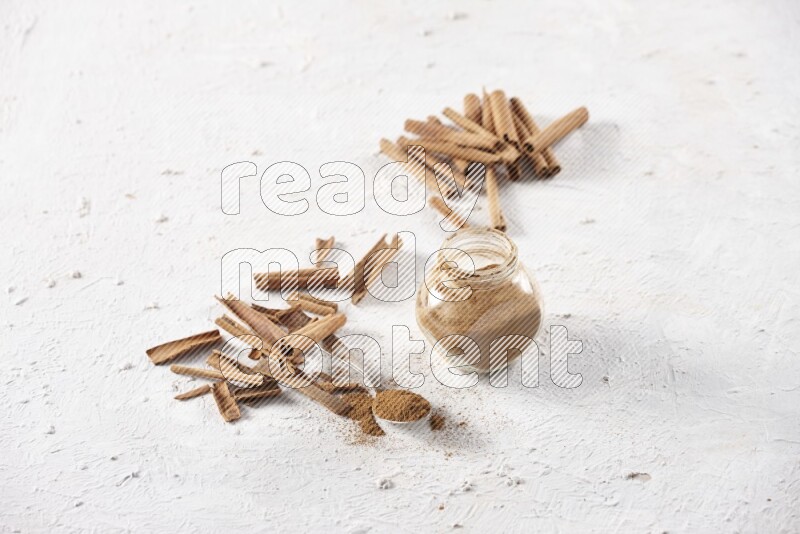 Herbal glass jar full cinnamon powder and a metal spoon surrounded by cinnamon sticks on a white background