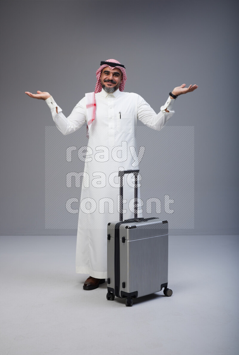 Saudi man wearing Thob and red Shomag standing holding Travel bag on Gray background