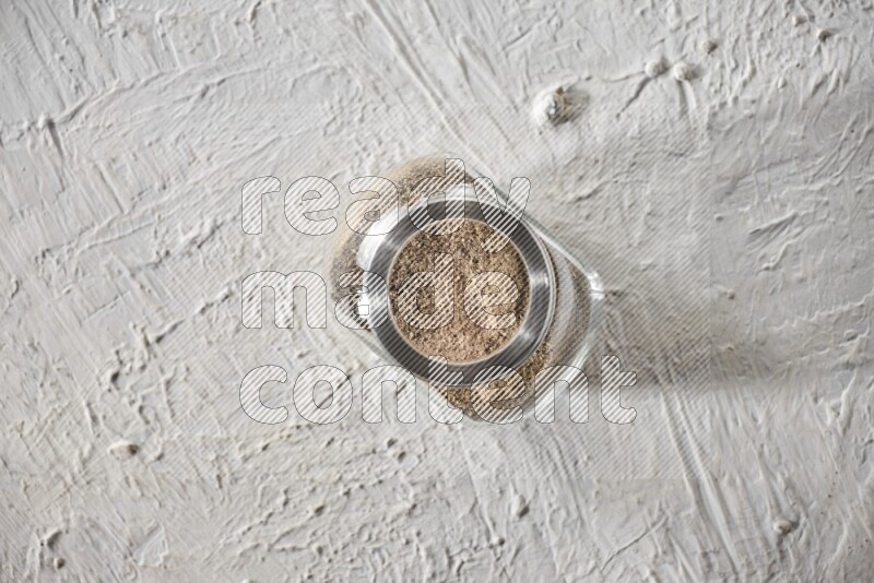 A glass spice jar full of powder black pepper on textured white flooring