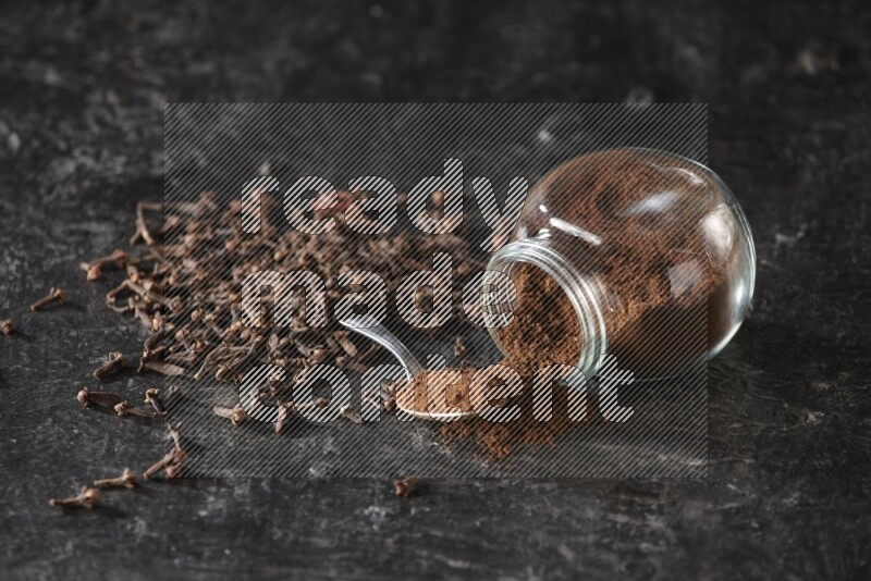 A flipped glass spice jar and a metal spoon full of cloves powder with cloves spread on textured black flooring