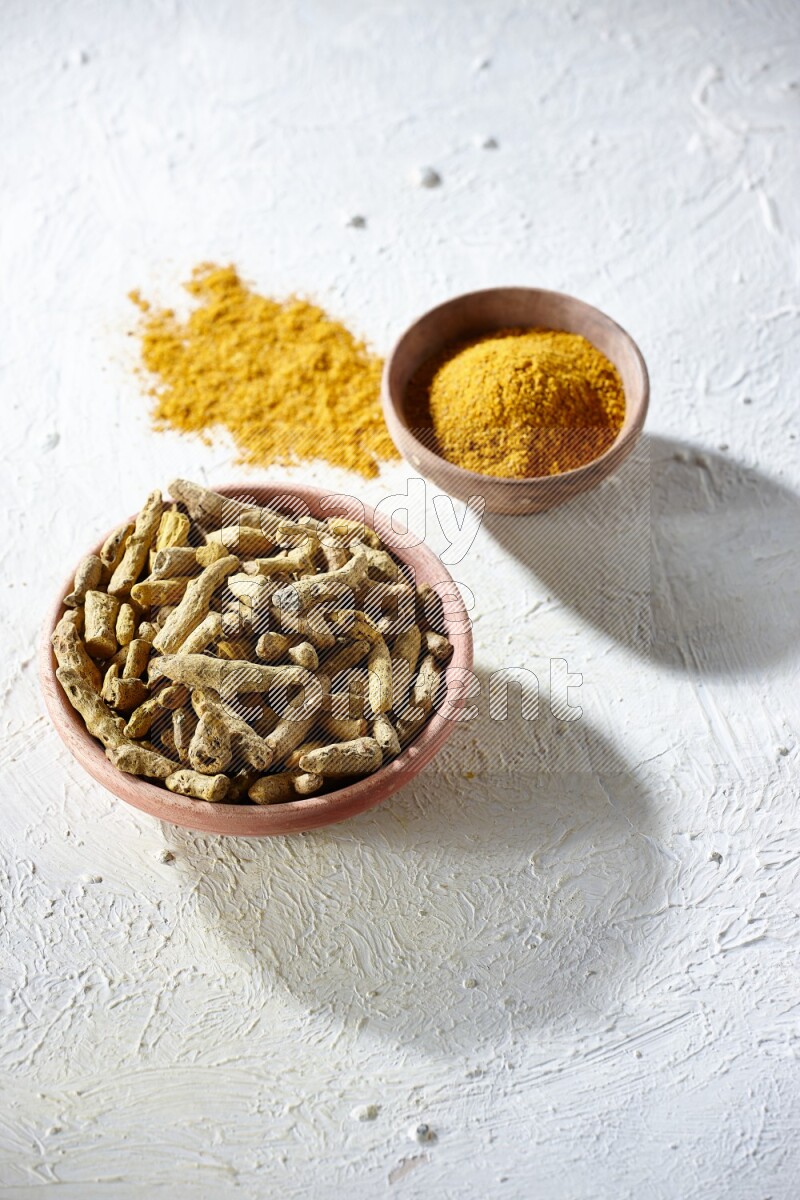 2 wooden bowls, one full of turmeric powder and the other full of dried turmeric whole fingers on a textured white flooring