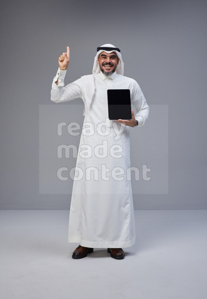 Saudi man Wearing Thob and white Shomag standing showing tablet to camera on Gray background