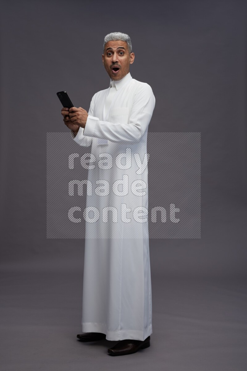 Saudi man wearing thob with neck strap employee badge standing texting on phone on gray background