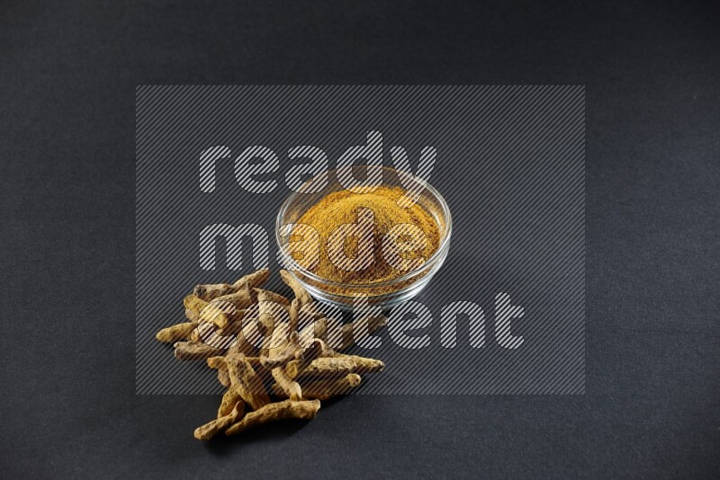 وعاء زجاجي ممتلئ ببودرة الكركم وأصابع الكركم المجففة بجانبه على خلفية سوداء