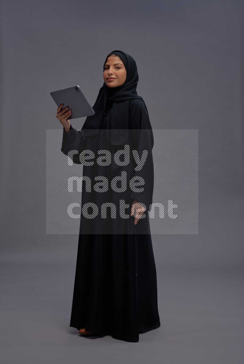 Saudi woman wearing Abaya standing working on tablet on gray background