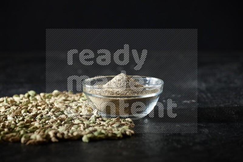 A glass bowl full of cardamom powder and cardamom seeds spreaded beneath the bowl on textured black flooring