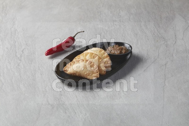 Three fried sambosas in an oval shaped black plate and a sauce in a black round ramekin on a gray background