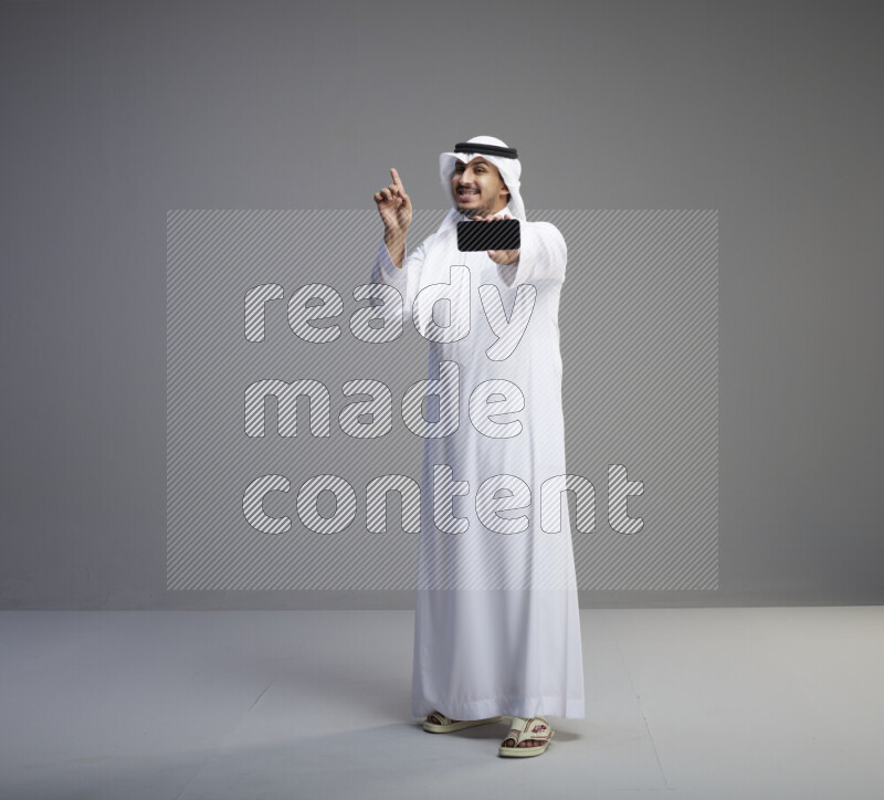 A Saudi man standing wearing thob and white shomag showing phone to camera on gray background