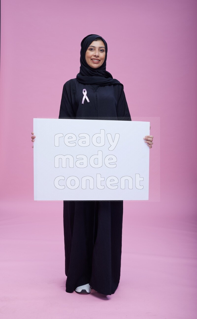 Saudi woman wearing pink ribbon on Abaya standing holding board on pink background
