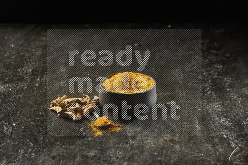 A pottery black bowl and a metal spoon full of turmeric powder with dried turmeric fingers on a textured black flooring