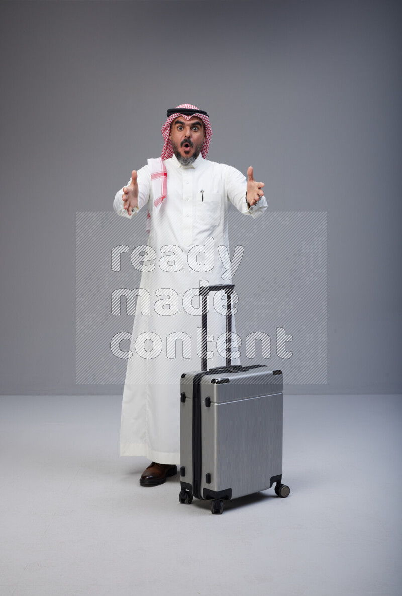 Saudi man wearing Thob and red Shomag standing holding Travel bag on Gray background