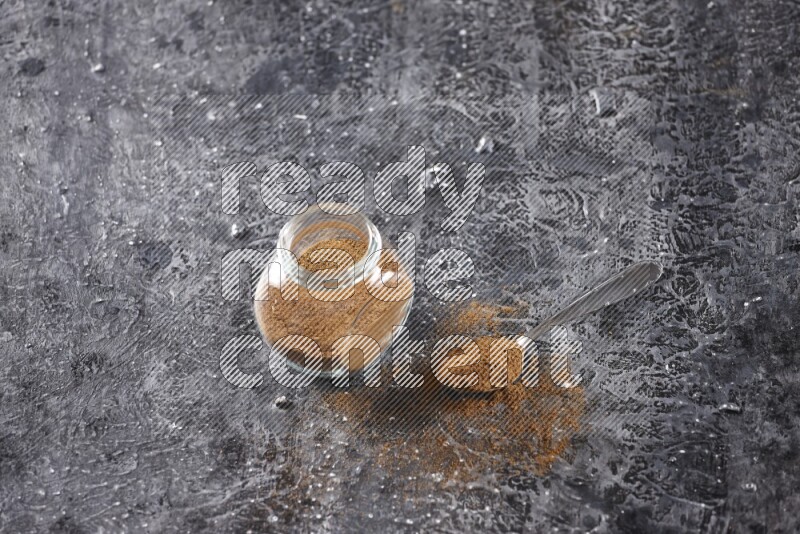 Herbal glass jar full of cinnamon powder and a metal spoon full of powder on textured black background