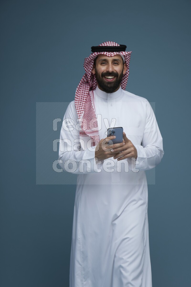 A Male posing with a phone in a blue background wearing Saudi Thob and Shomag