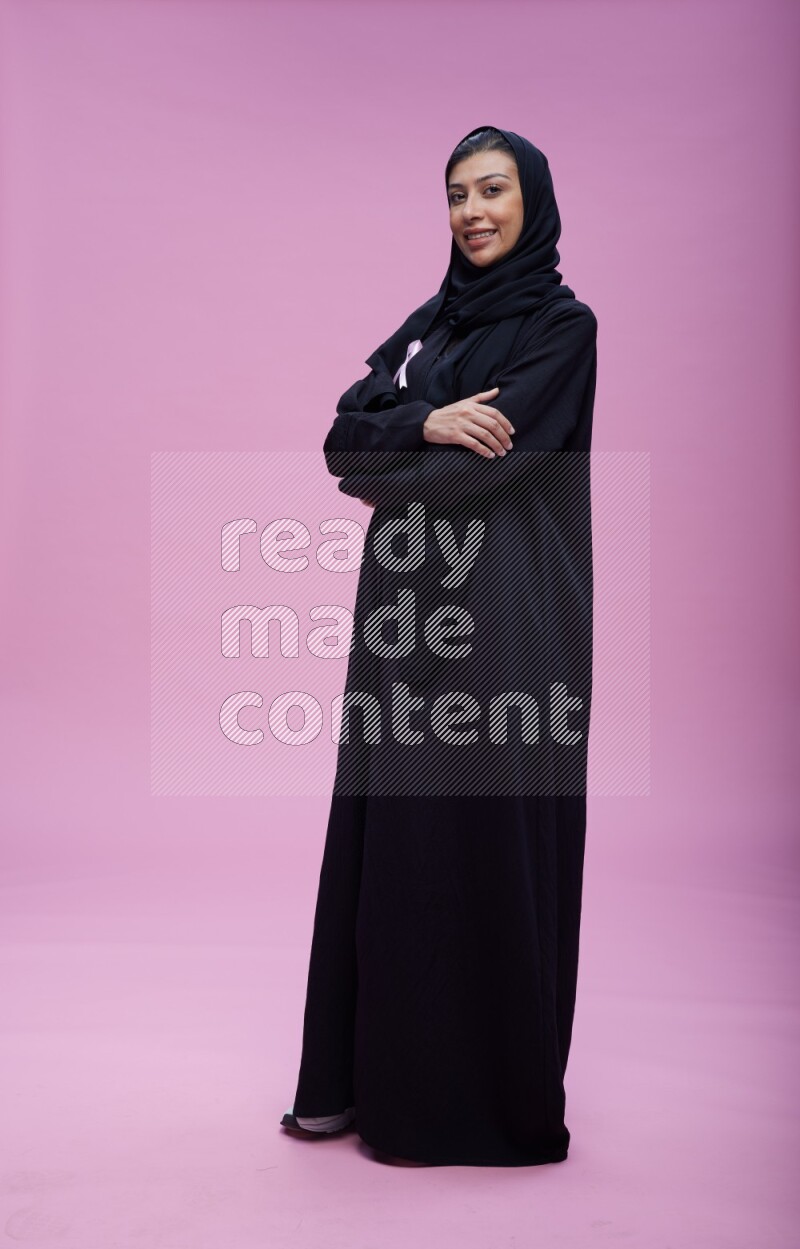 Saudi woman wearing pink ribbon on Abaya standing crossed arms on pink background