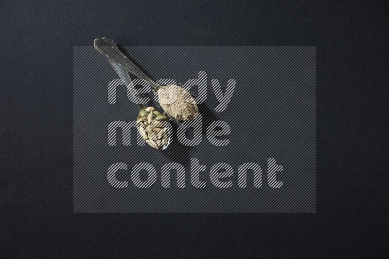 ملعقتان معدنيتان ممتلئتان ببودرة الهيل وحبوب الهيل على خلفية سوداء