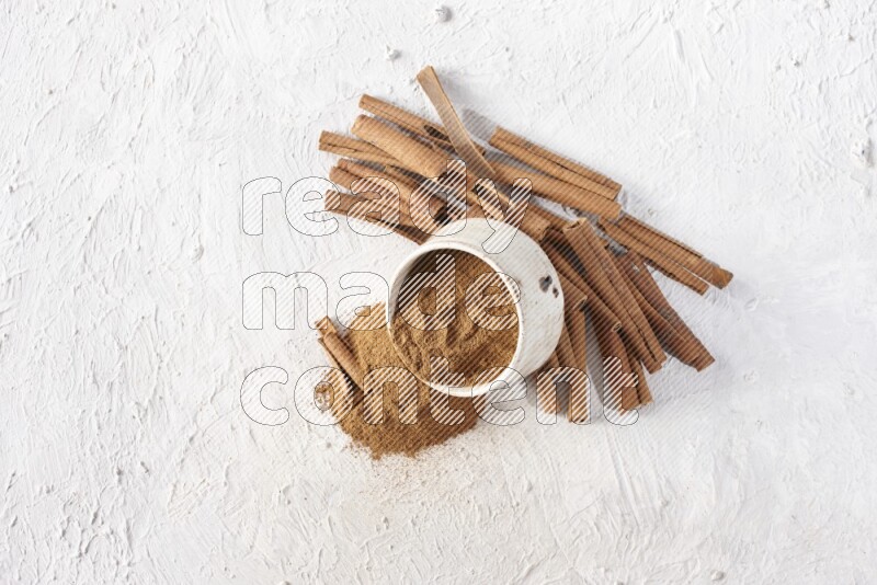 Ceramic beige bowl over filled with cinnamon powder and cinnamon sticks around the bowl on a textured white background in different angles