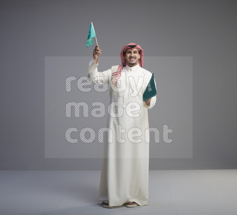 A Saudi man standing wearing thob and red shomag raising small Saudi flag on gray background