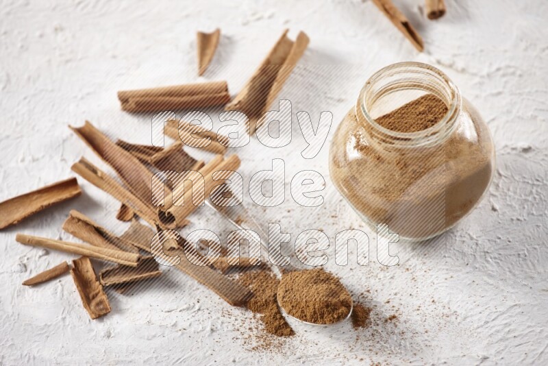 Herbal glass jar full cinnamon powder and a metal spoon surrounded by cinnamon sticks on a white background