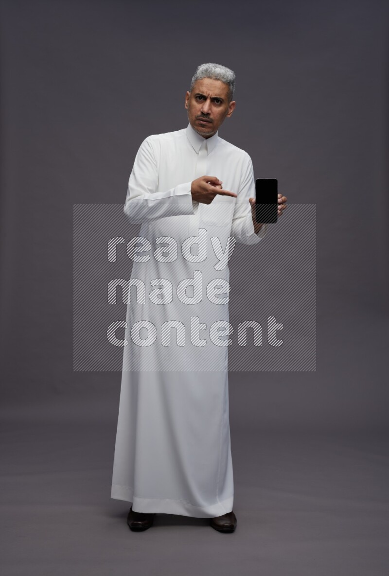 Saudi man wearing thob standing showing phone to camera on gray background