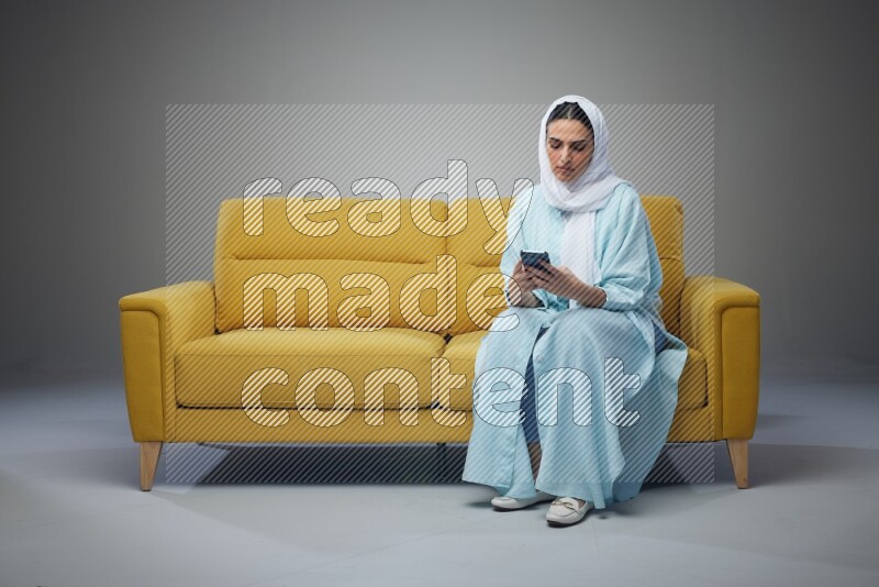 A Saudi woman wearing a light blue Abaya and white head scarf sitting on a yellow sofa and using her phone eye level on a grey background