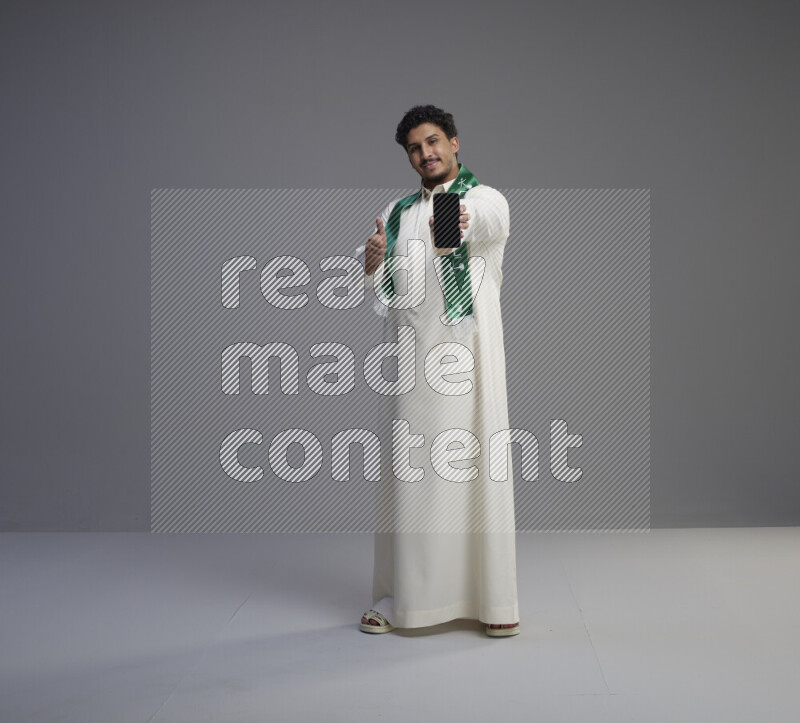 A Saudi man standing wearing thob and Saudi flag scarf showing phone to camera on gray background