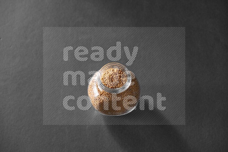 A glass spice jar full of mustard seeds on black flooring