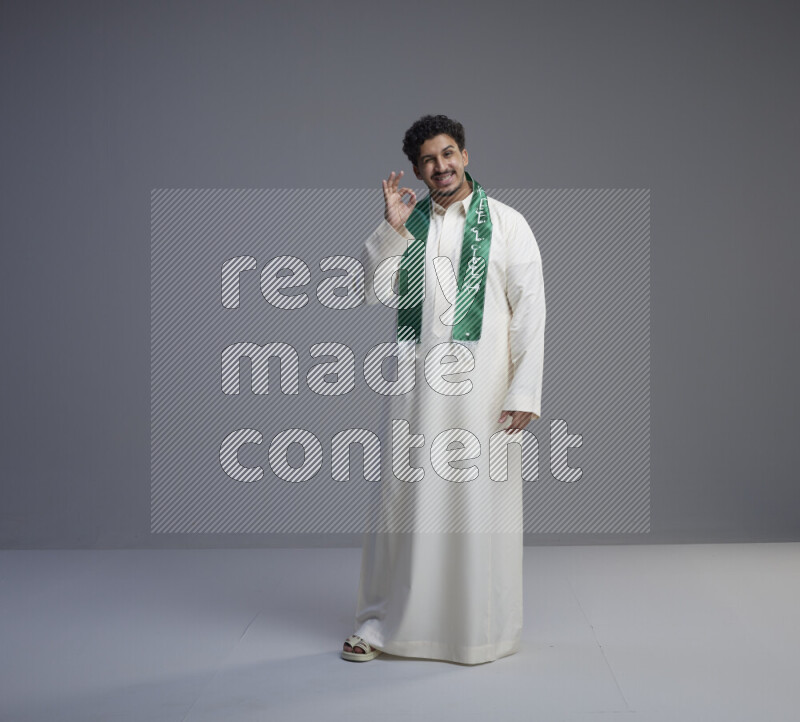 A Saudi man standing wearing thob and Saudi flag scarf on gray background