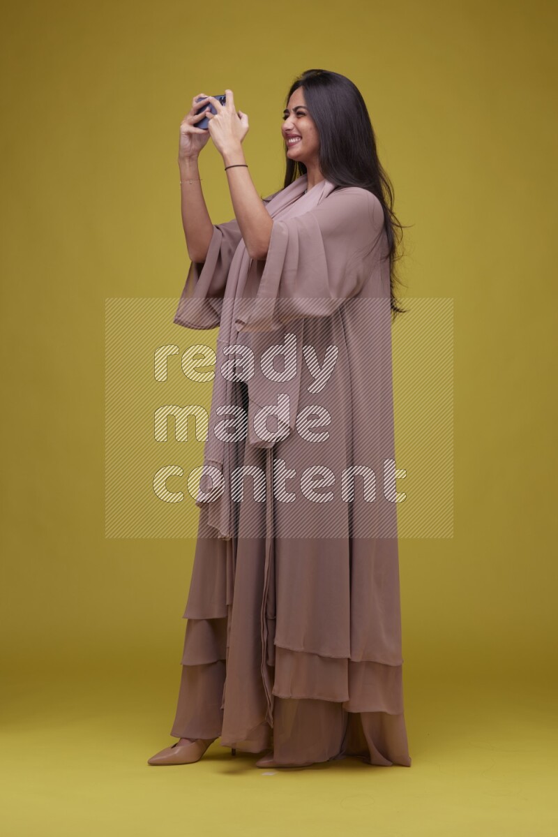 A woman Playing a Game on her smartphone on a Yellow Background wearing Brown Abaya