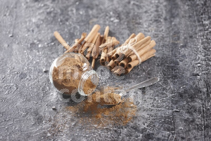 Herbal glass jar full cinnamon powder flipped and a metal spoon full of powder, cinnamon sticks stacked and bounded in the back on textured black background in different angles